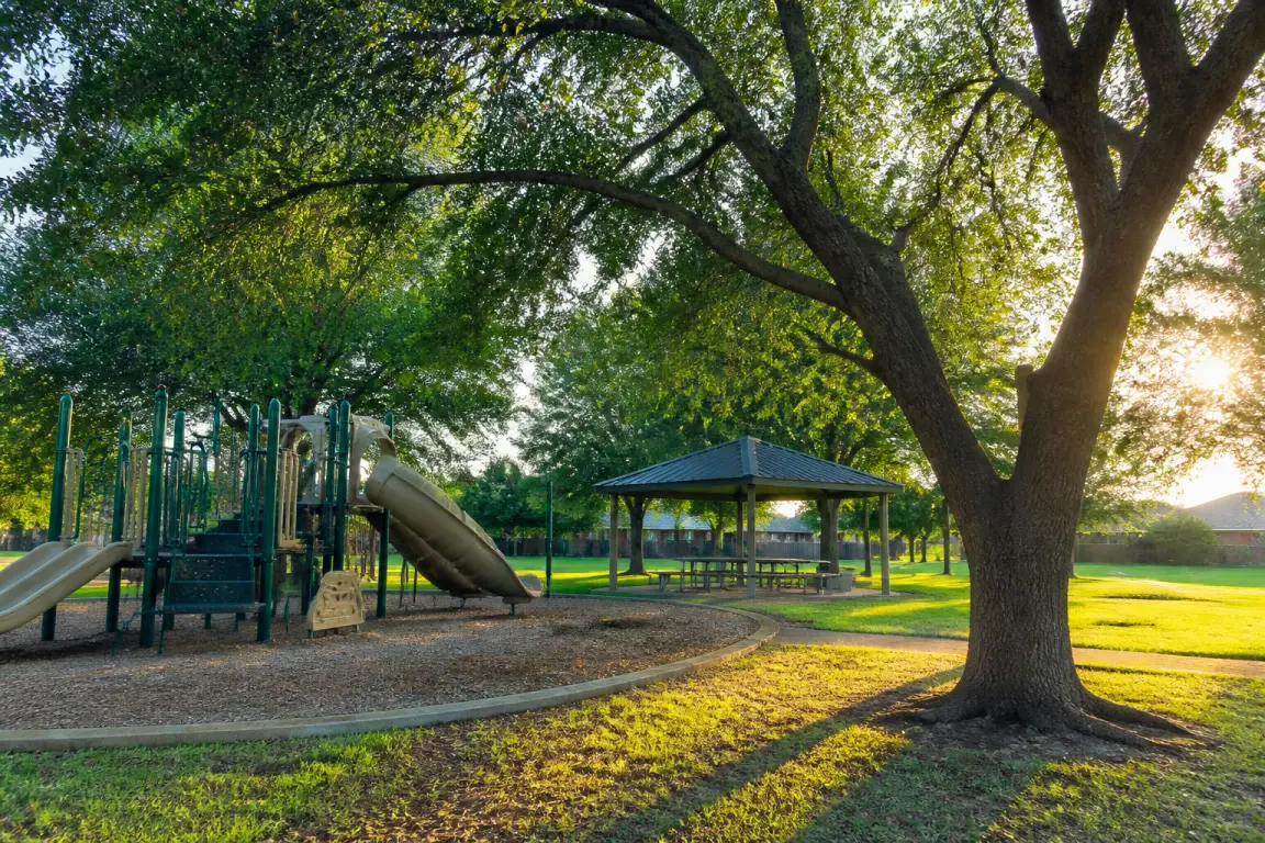 A neighborhood park with a playground and a covered picnic pavilion beneath a large oak tree.