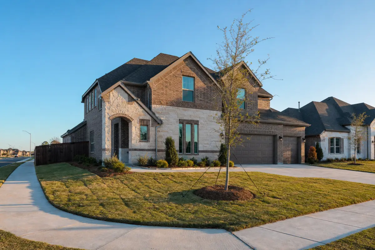 A two-story brick and stone home on a corner lot with new sod and a young staked tree.