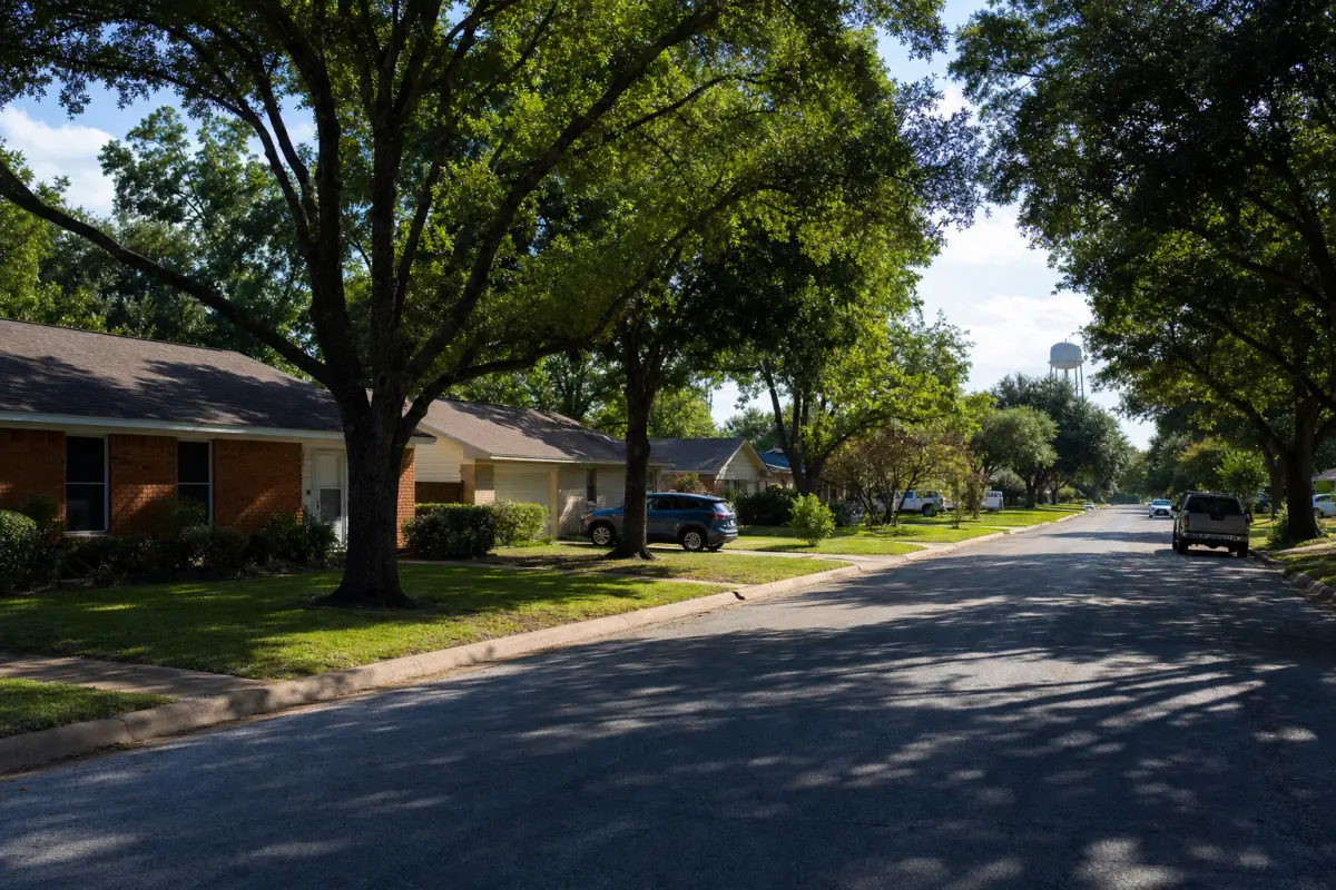A small-town residential street of single-story homes under large shade trees.