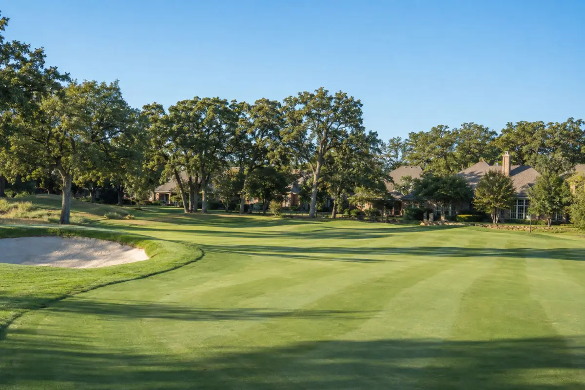 A golf fairway with a sand bunker and mature trees, with the rooflines of homes beyond.